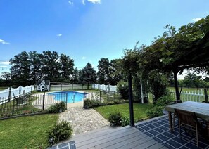 Un pati amb piscina, terrassa de fusta, porxo cobert i tanca blanca.