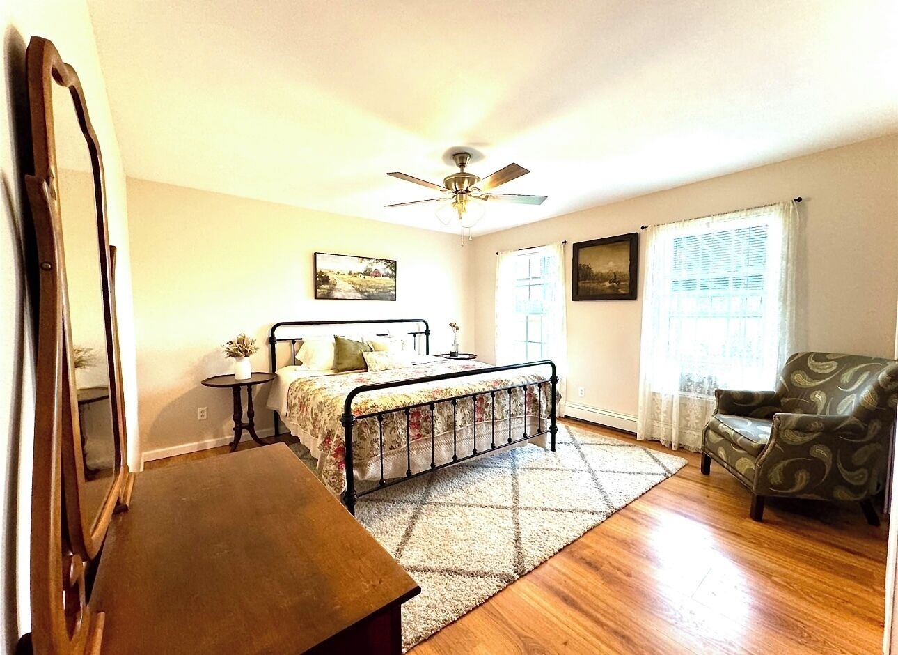 Une chambre à coucher avec un lit, une chaise, une table, un ventilateur de plafond et une fenêtre avec des rideaux.