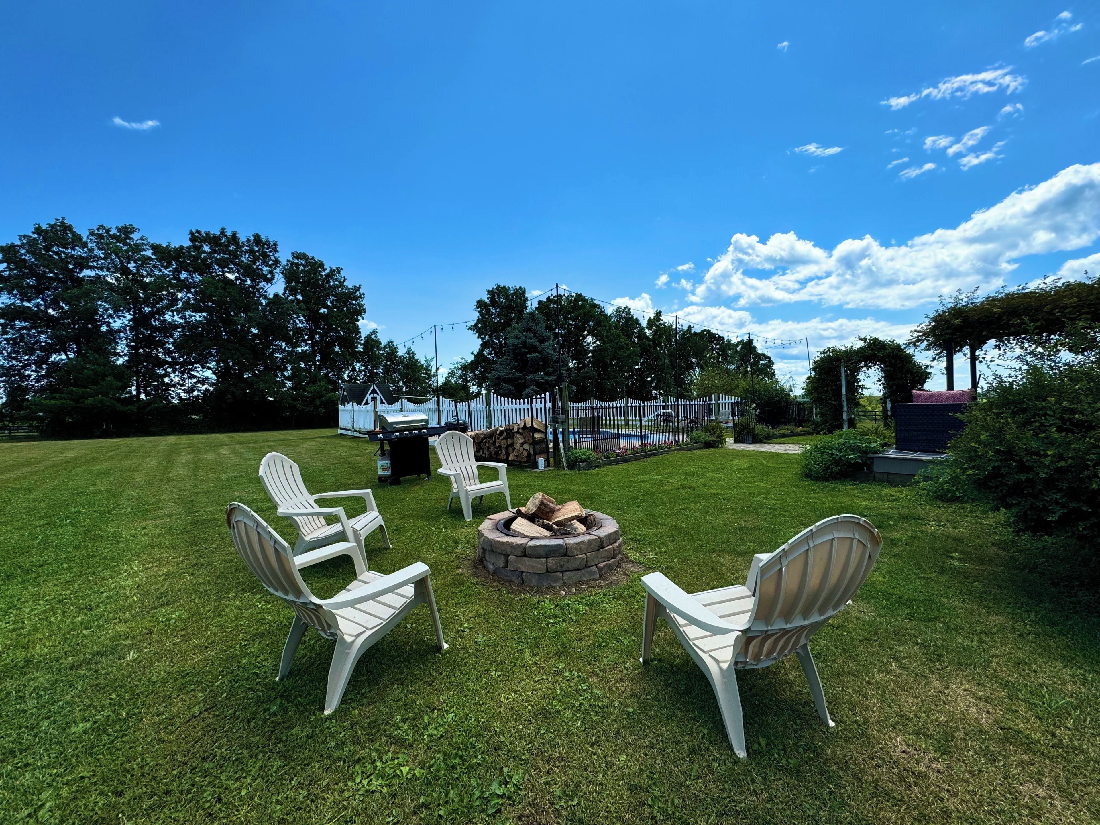 A grassy outdoor area with a fire pit, white chairs, and a white fence.