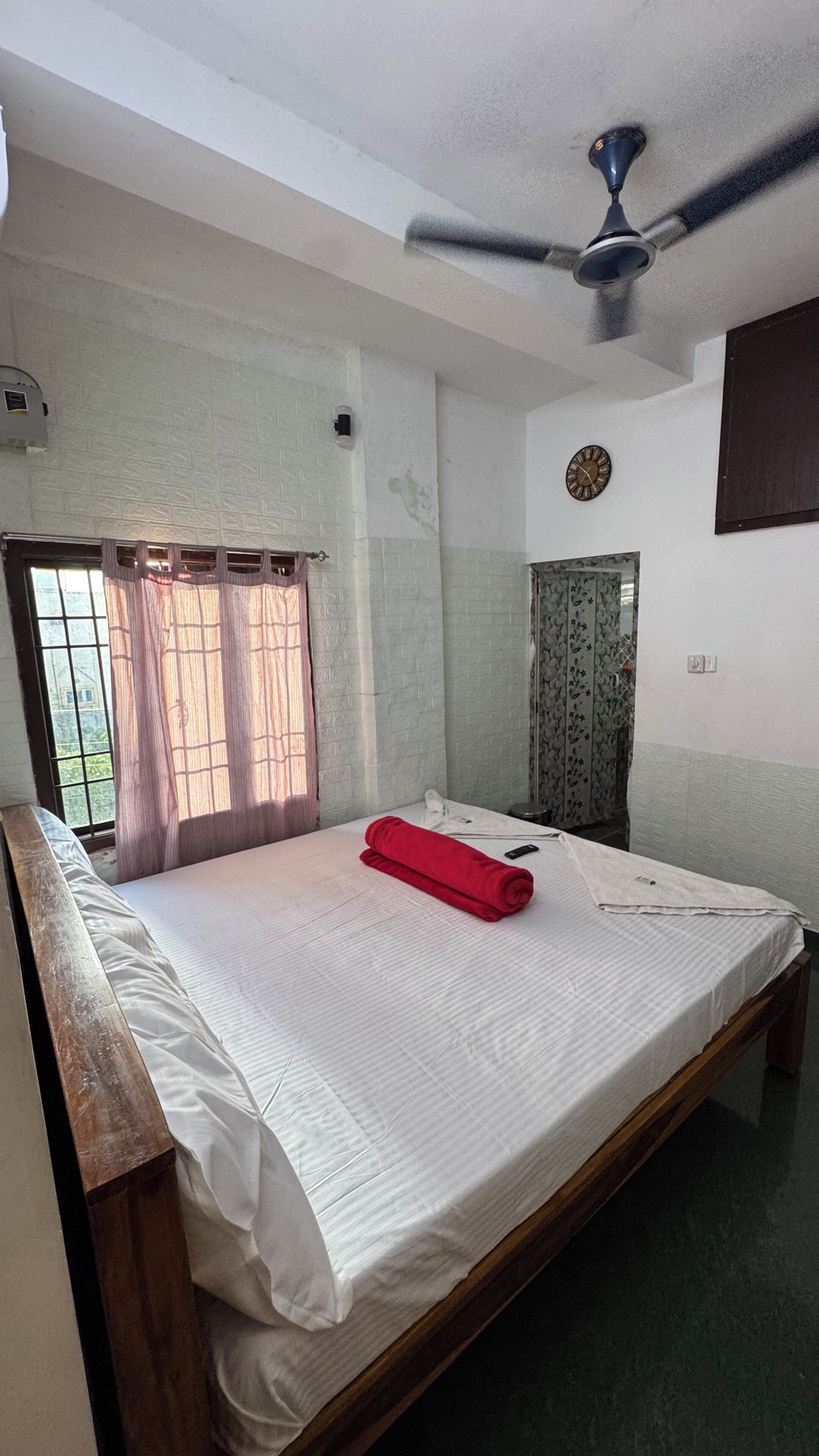 A room with a bed, a ceiling fan, a window with curtains, a clock on the wall, and a door with a patterned curtain.