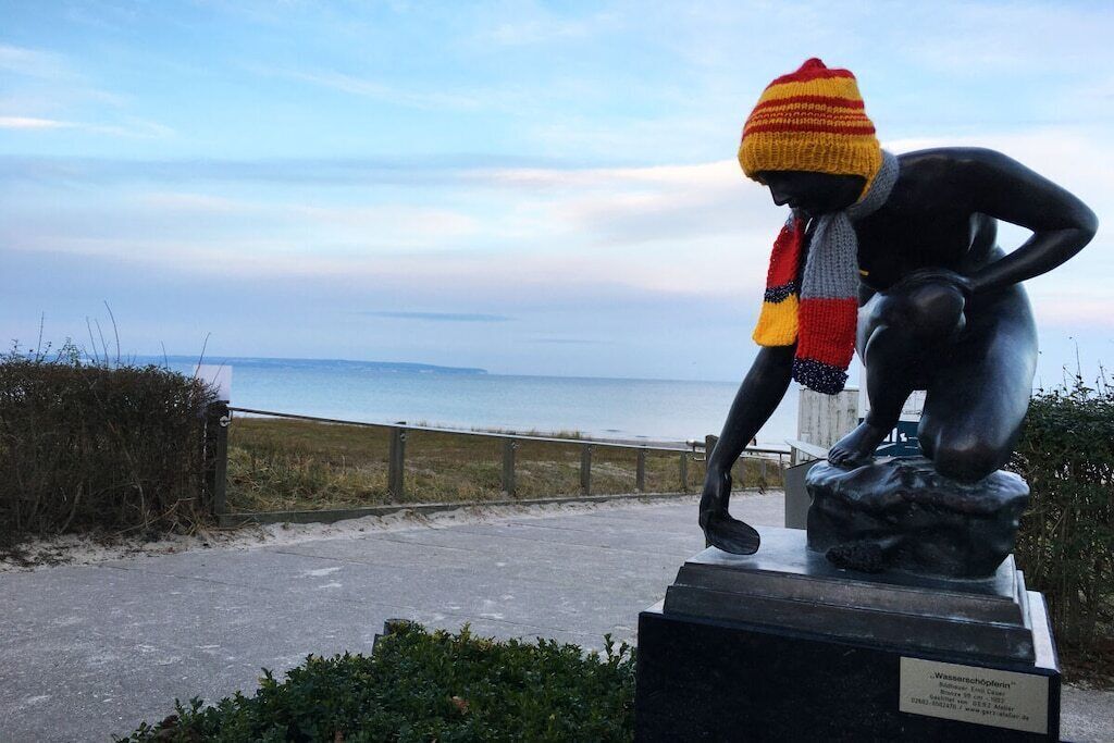 Une sculpture représentant une figure coiffée d’un bonnet et portant une écharpe tricotés, placée sur un piédestal agrémenté d’une plaque, et dominant un sentier côtier.