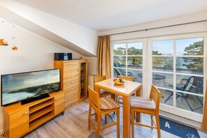 A room with a TV on a wooden stand, a dining table with chairs, and a view through large windows. - Seaside joy at the Baltic Sea House (Binz)