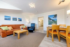 A modern living room with a sofa, coffee table, and dining area. There are wall-mounted lights and artwork. - Seaside joy at the Baltic Sea House (Binz)