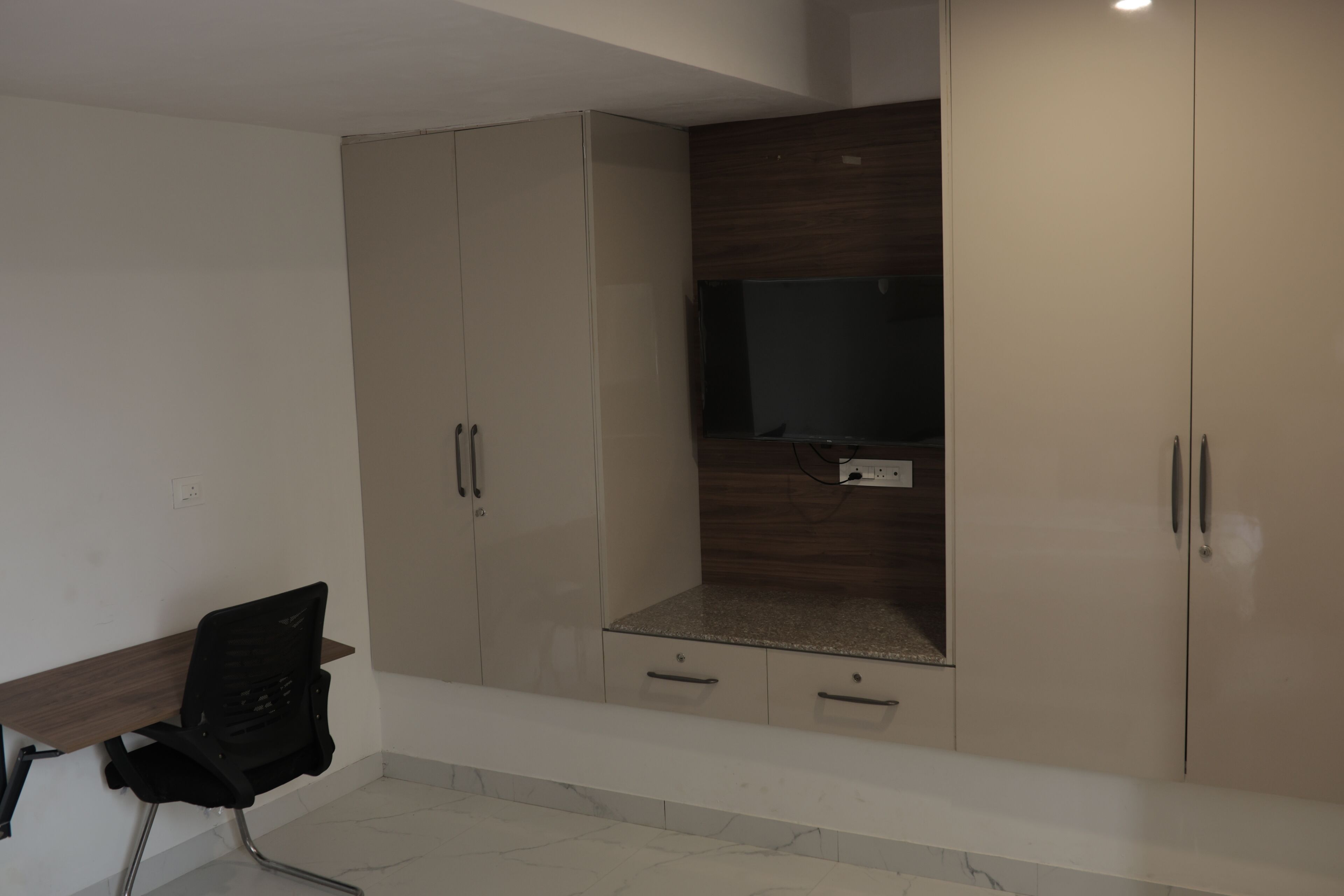 A room with a wardrobe, a television mounted on the wall, and a desk with a chair.