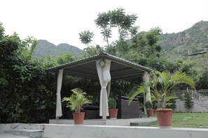 A covered outdoor seating area with a roof, surrounded by potted plants and a mountainous backdrop.