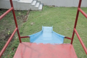 A playground slide with a red and blue structure, a grassy area, and a concrete staircase.