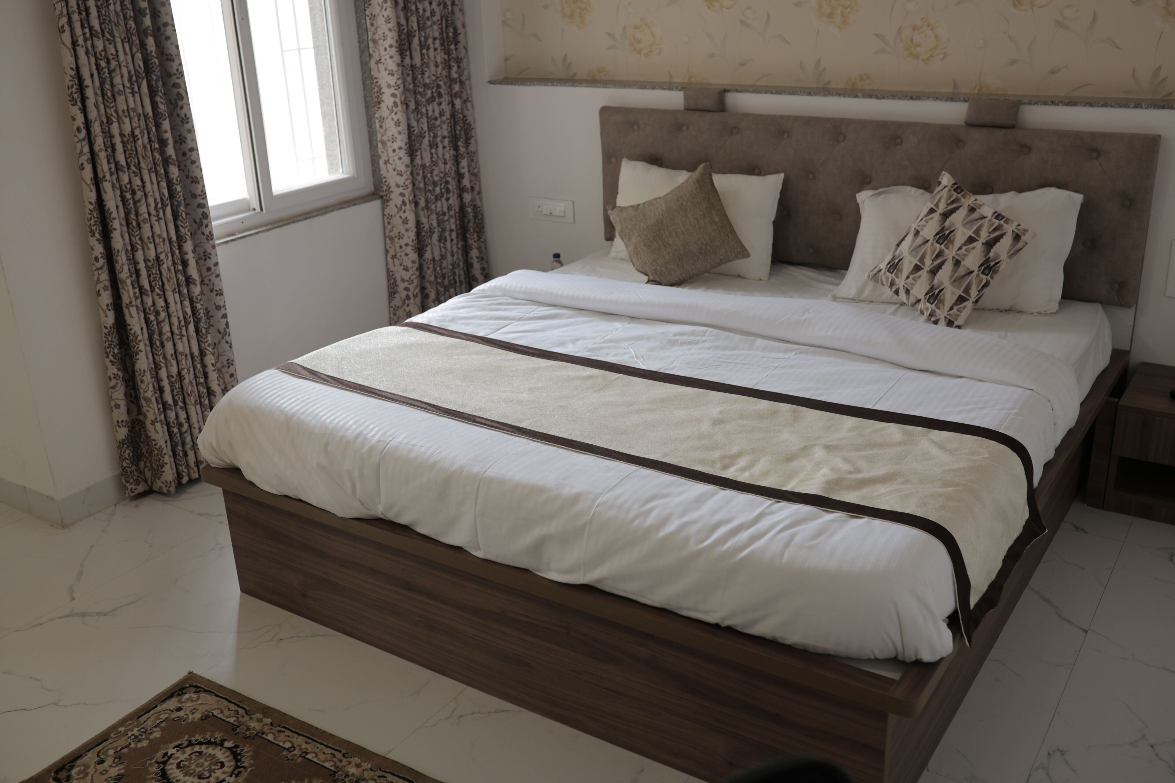 Een slaapkamer met een bed, gordijnen, een hoofdeinde en een kamerbreed tapijt met een patroon.