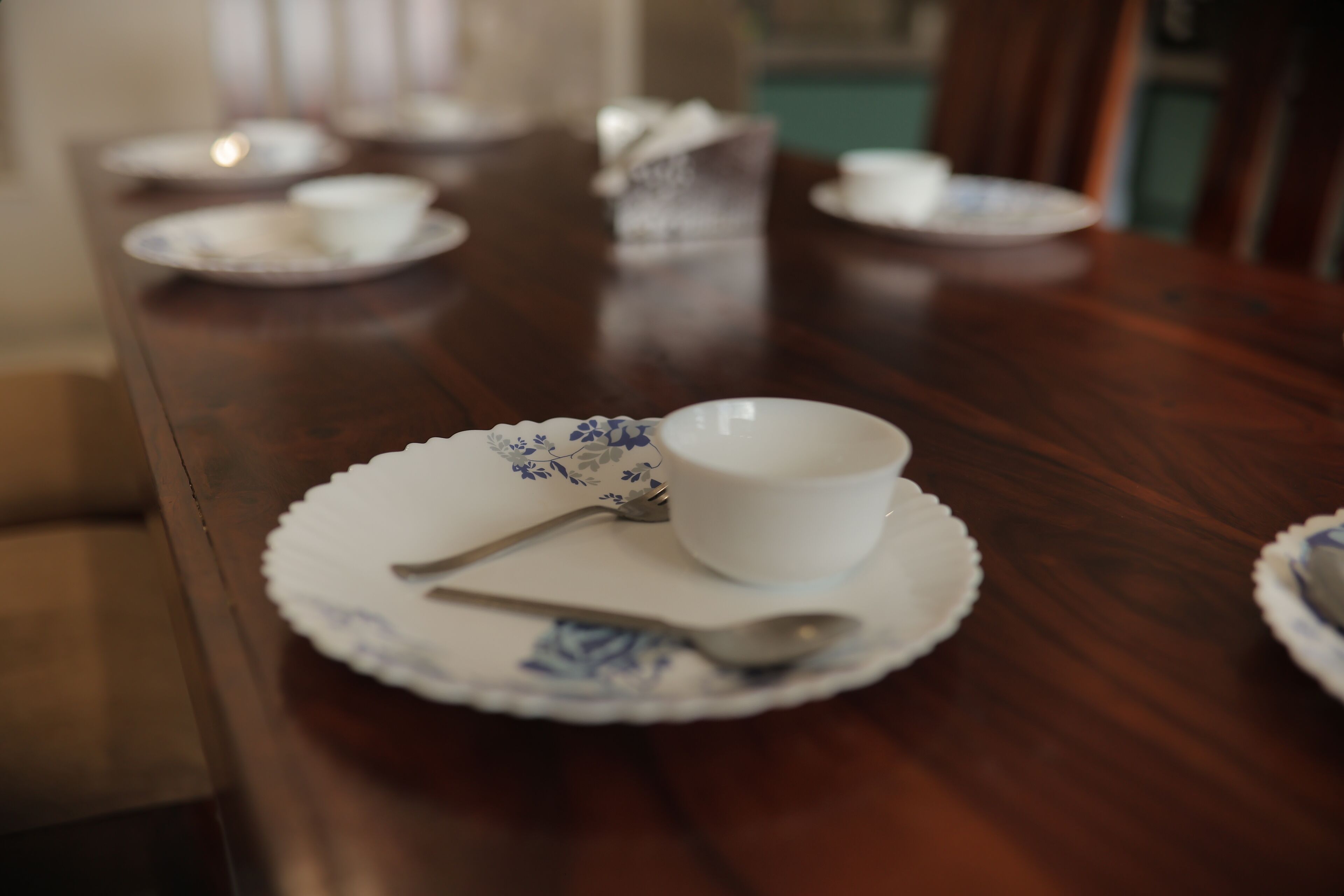 A dining table with a white bowl and spoon on a floral plate, set in a room with a wooden table and chairs.