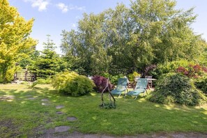 Un jardin avec un sentier, des chaises vertes et divers arbustes.