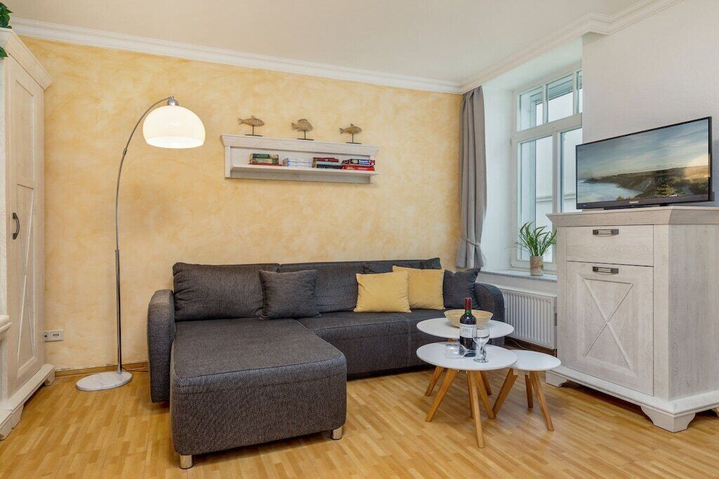 A living room with a grey sofa, a floor lamp, a flat-screen TV, and a small white table with a bottle and glass.