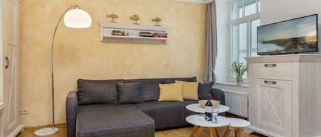 A living room with a grey sofa, a floor lamp, a flat-screen TV, and a small white table with a bottle and glass.