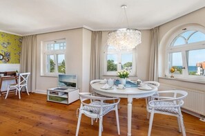A dining area with a round table, white chairs, a chandelier, and large windows with curtains. - Holiday apartment in Binz with swimming pool (Binz)