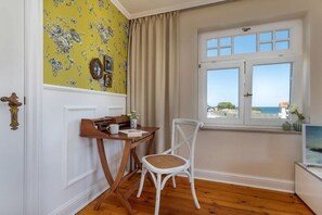 A room with a wooden desk, a white chair, a window with a view of the sea, and a yellow floral wallpaper. - Holiday apartment in Binz with swimming pool (Binz)