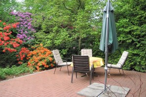 An outdoor patio with a table and chairs, surrounded by lush greenery and flowering plants. - Hof Freitag Geestequelle (Hipstedt)