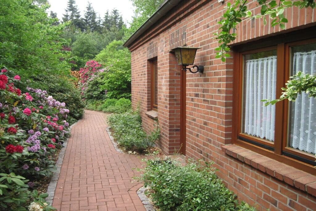 A brick building with a brick pathway leading to a door, surrounded by lush greenery and flowering plants.