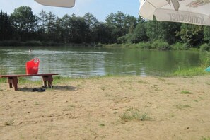 A sandy beach with a bench and a red bucket, a calm lake with swimmers, and a white umbrella. - Hof Freitag Geestequelle (Hipstedt)