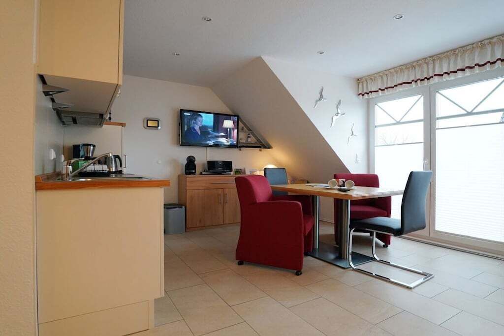 A modern kitchen with a dining area, a TV mounted on the wall, and a large window with curtains.