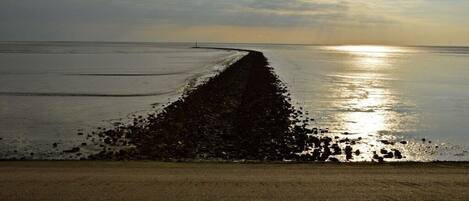 A beach with a long, narrow path leading to the water, a clear sky, and the sun low on the horizon.