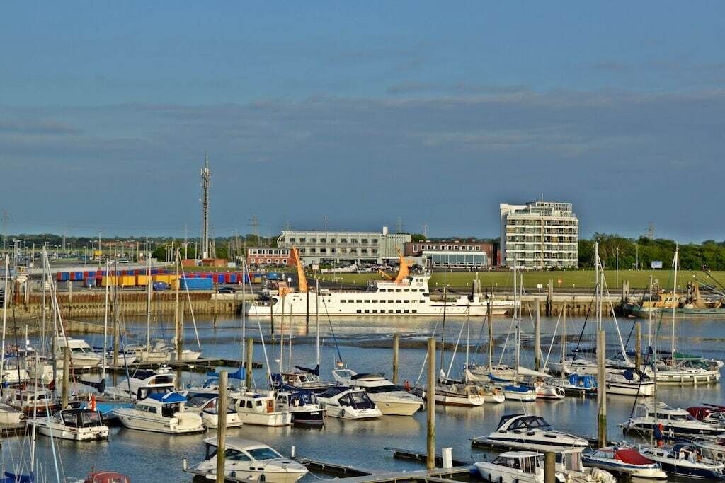 A marina with multiple boats docked, a large cruise ship, and a modern building in the background.