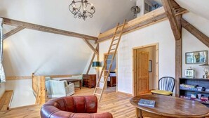 A loft bedroom with exposed wooden beams, a loft bed, a leather armchair, a wooden dining table, and a ladder.
