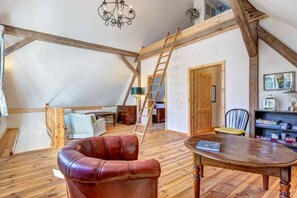 A loft bedroom with exposed wooden beams, a loft bed, a leather armchair, a wooden dining table, and a ladder.