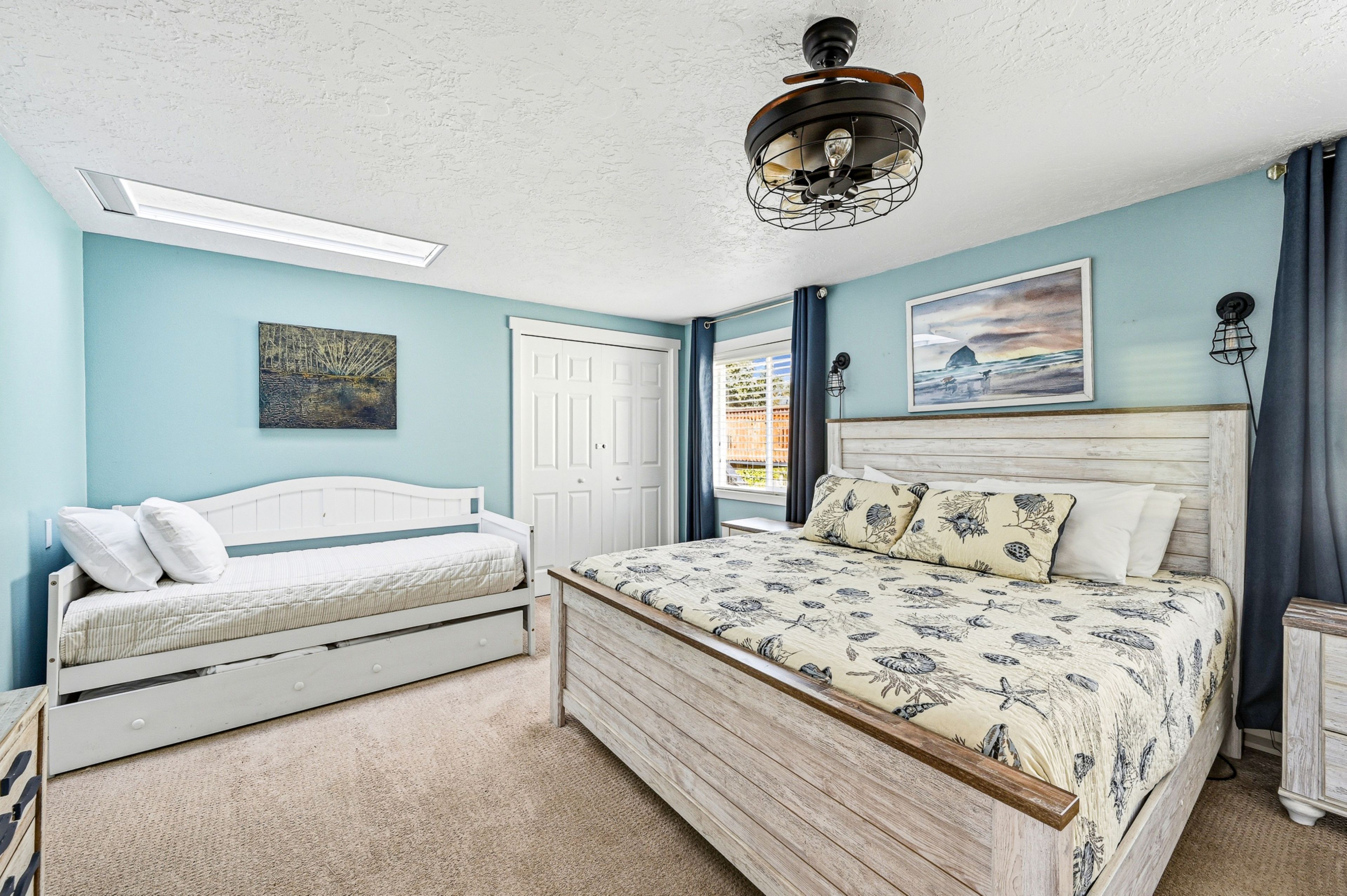 A bedroom with a bed, a smaller bed, a ceiling fan, a painting, and a door.