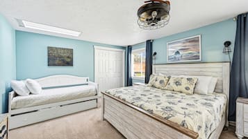 A bedroom with a bed, a smaller bed, a ceiling fan, a painting, and a door.