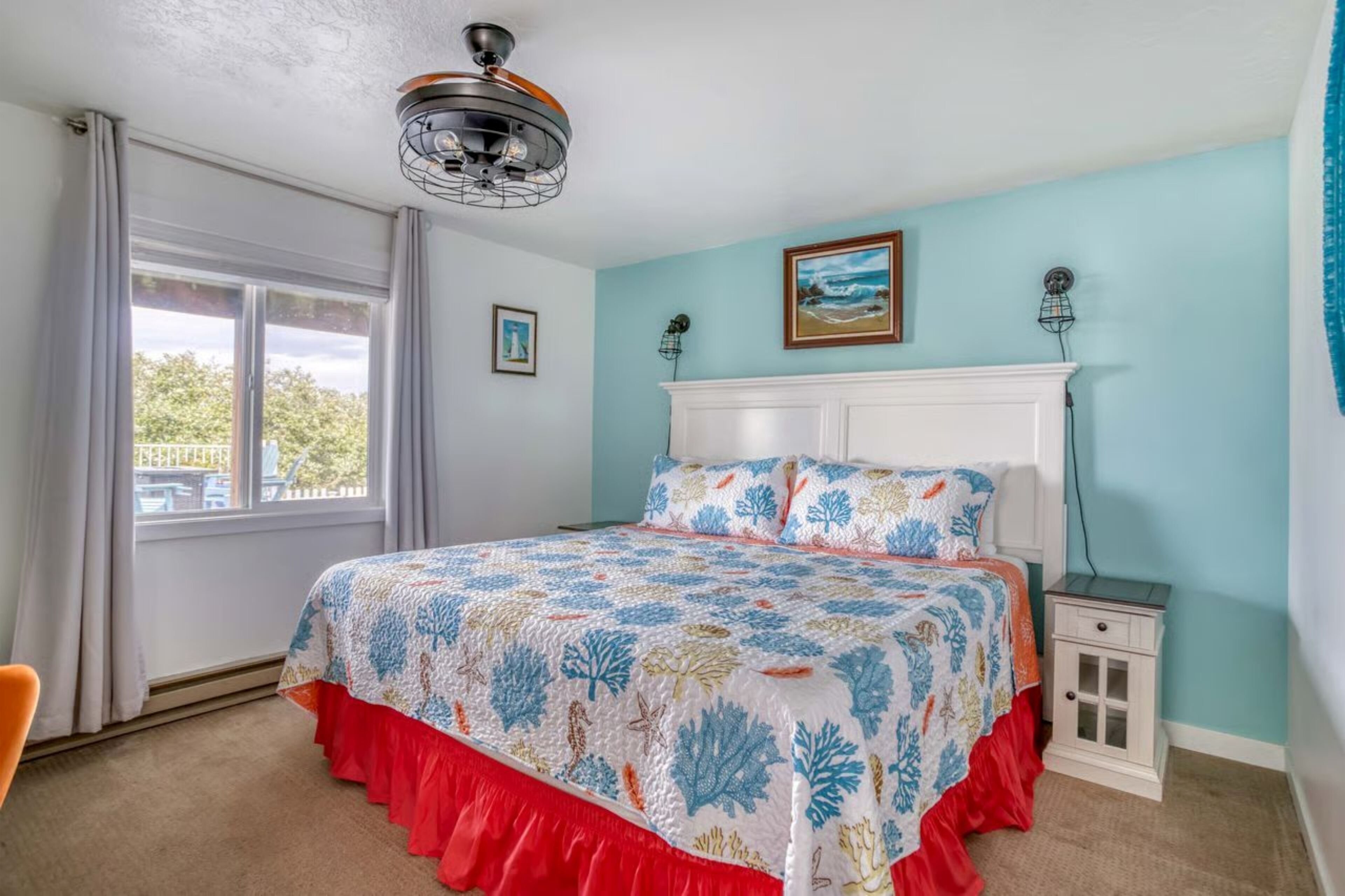 Una camera da letto con un letto, un comodino, una finestra con tende, una ventola a soffitto e un quadro appeso al muro.