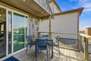 A wooden deck with a table and chairs, a glass door leading to a room, and a building with siding and a red roof. - Lincoln City Amazing View Retreat: Hot Tub, Arcade Room, Steps to Beach (Lincoln City)