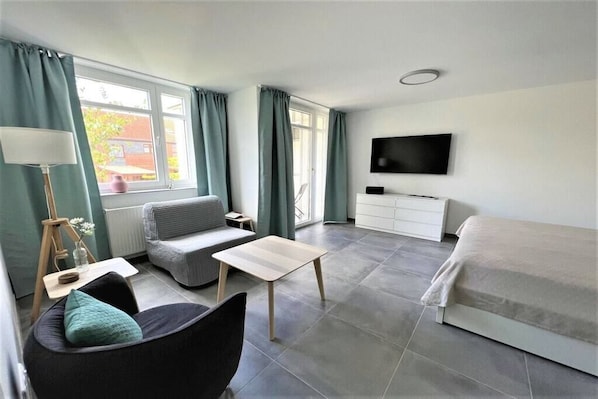 A modern room with a bed, a sofa, a small table, a TV, and a window with curtains. - Holiday apartment with 1 bedroom (Neubrandeburg)
