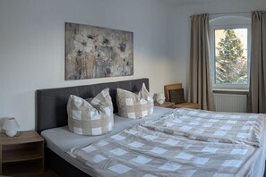 Ein Schlafzimmer mit Bett, Kissen, einem Nachttisch, einer Lampe, einem Bild an der Wand und einem Fenster mit Vorhängen.