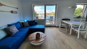 Un saló amb un sofà blau, una taula de centre rodona, una taula de dinar amb cadires i una vista del port esportiu a través d'una porta de vidre.