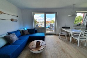 Ein Wohnzimmer mit blauem Sofa, einem runden Couchtisch, einem Esstisch mit Stühlen und Blick auf einen Yachthafen durch eine Glastür.