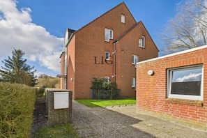 A red brick building with the letters 'LIG' on the facade, a gravel path, and a small grassy area. - Siefert holiday apartment (Langeoog)