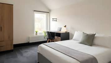A bedroom with a bed, a desk with a laptop, a radiator, and a window.