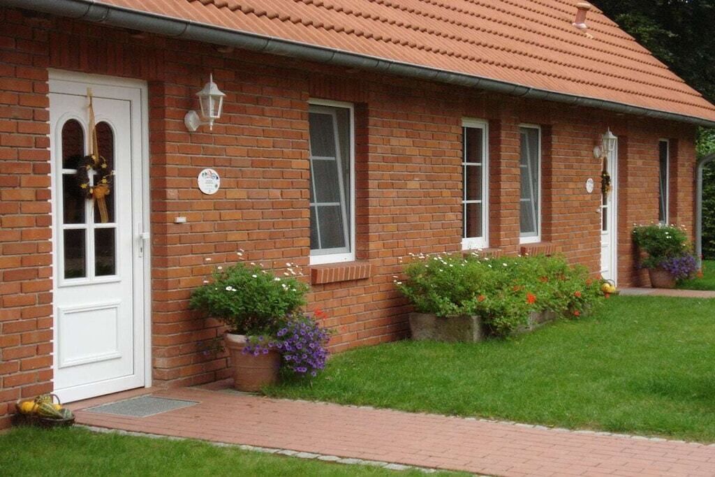 Een bakstenen huis met een witte deur, ramen en een pannendak. Er staan bloempotten met planten en een geplaveid pad.