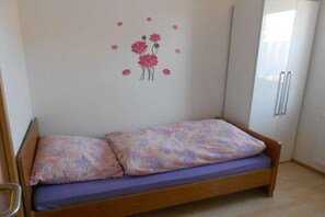 A single bed with a floral-patterned bedspread and pillows, a wooden headboard, and a wall decoration of pink flowers. - Sonja's holiday apartment (Großaitingen)