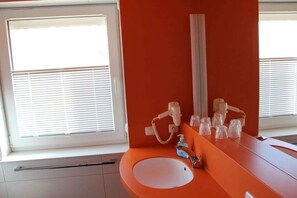 Une salle de bain avec un lavabo blanc, un plan de toilette orange et une fenêtre avec des stores.