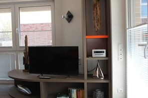 A living room with a flat-screen TV on a stand, a bookshelf with books and decorative items, a small table with a lamp, and a window with blinds. - 10 in the Palais am Meer II (Cuxhaven-Duhnen)