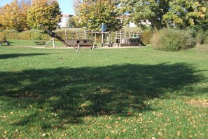 Een speeltuin met diverse speeltoestellen, waaronder een glijbaan en klimrekken, gelegen in een grasveld met bomen en een bank op de achtergrond.