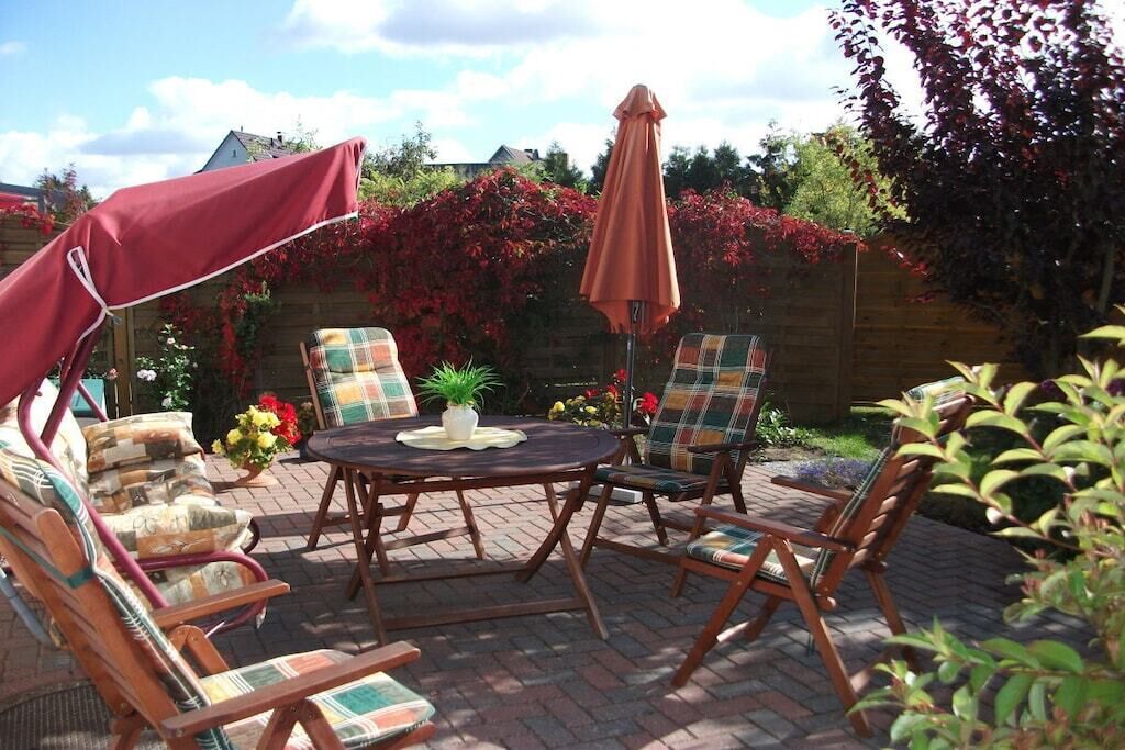 Un patio exterior con una mesa redonda y cuatro sillas, rodeado por una cerca de madera y plantas con flores.