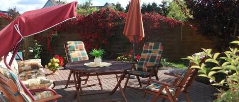 An outdoor patio with a round table and four chairs, surrounded by a wooden fence and flowering plants.