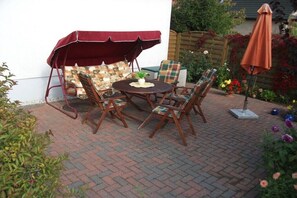Eine Terrasse mit einem runden Tisch, vier Stühlen, einem roten Sonnensegel und einem Blumentopf.