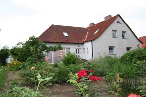 A two-story house with a sloped roof and a garden with various plants and flowers. - Holiday apartment with 1 bedroom (Waren (Müritz))