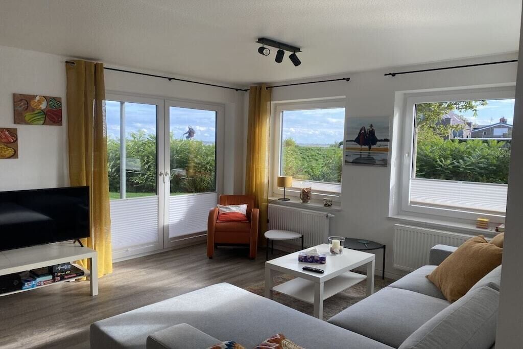 Un salon moderne avec un téléviseur à écran plat, un canapé, une table basse et un fauteuil. De grandes fenêtres sont présentes, avec des rideaux, un radiateur et une vue sur la verdure à l’extérieur.