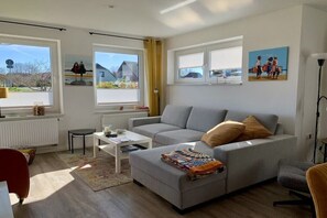 A living room with a grey sofa, a coffee table, and a patterned throw. There are two windows with blinds, a radiator, and a lamp. - Holiday apartment with 1 bedroom (Fehmarn)