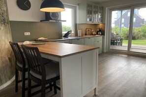A modern kitchen with a central island, dark chairs, and a view through a glass door to the outdoors. - Holiday apartment with 1 bedroom (Fehmarn)