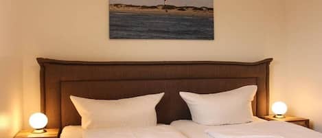 A neatly made bed with white linens and pillows, a headboard, and a nightstand with a lamp.