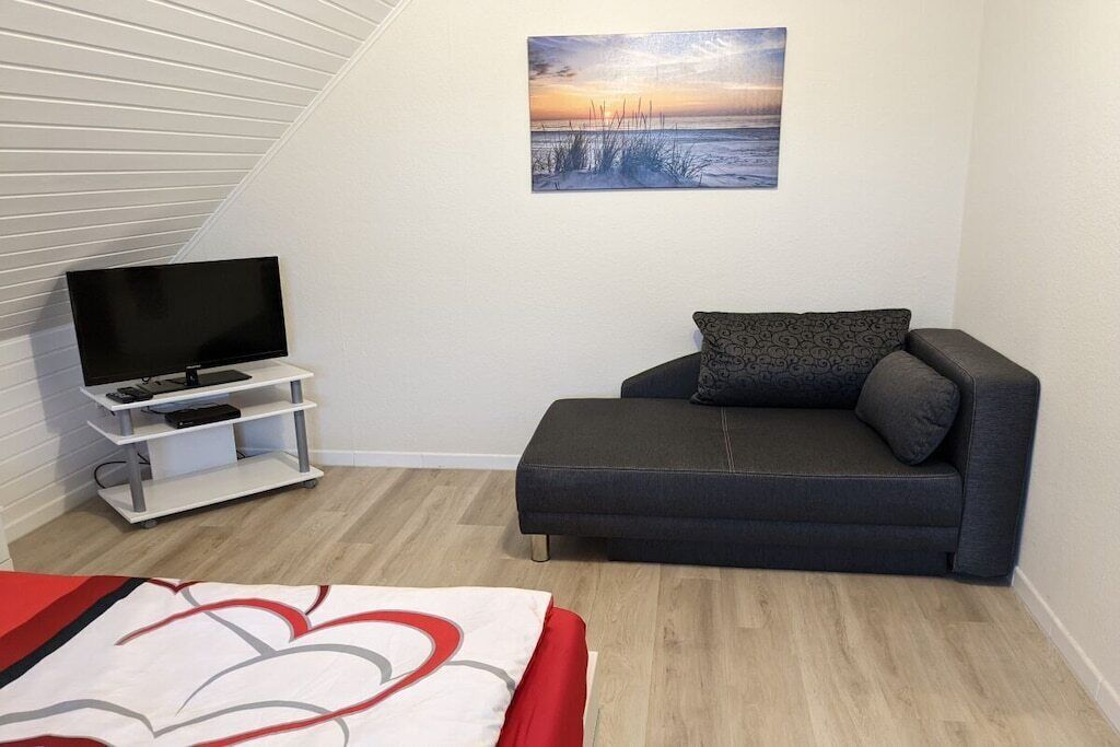 Une pièce avec un téléviseur à écran plat posé sur un meuble blanc, un canapé gris agrémenté d’un coussin à motifs et un tableau représentant un coucher de soleil accroché au mur.