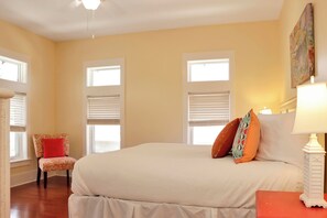 Un dormitorio con una cama grande, dos ventanas con persianas, una silla con un cojín rojo y un cuadro en la pared.
